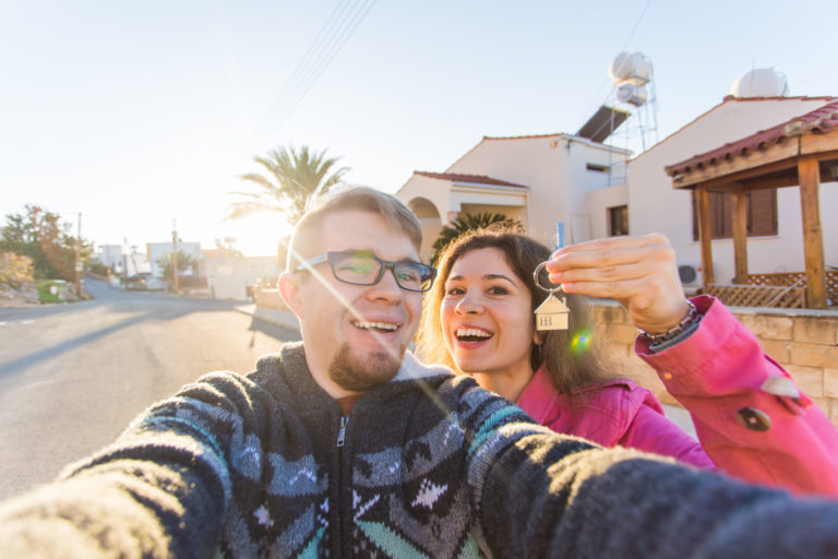 Happy young laughing cheerful couple man and woman handing their new home keys in front of a house